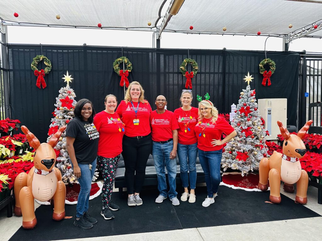 Six people, true Community Heroes, stand smiling in front of white Christmas trees, wreaths, red poinsettias, and inflatable reindeer Five wear matching red shirts and one a black shirt, creating a cheerful and festive scene
