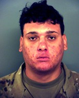 A young man with short, dark curly hair wearing a camouflage patterned shirt stares directly at the camera with a neutral expression against a plain background