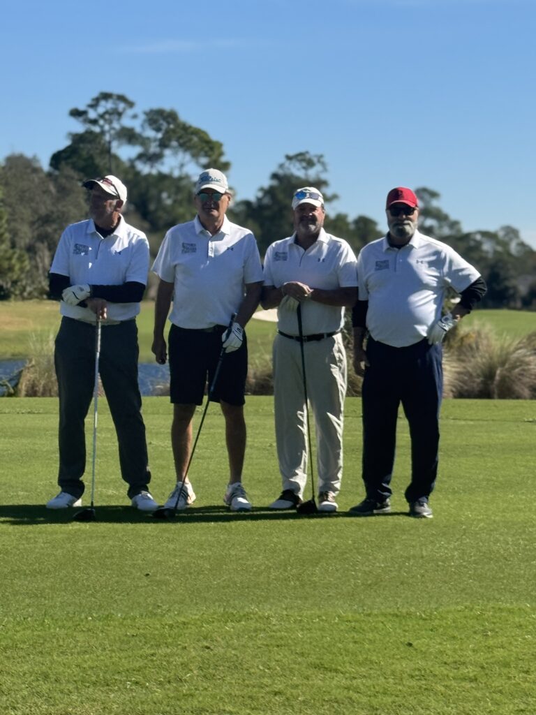 Four men stand on a golf course, wearing white shirts and holding golf clubs They pose together on the green, with trees, blue sky, and water in the background