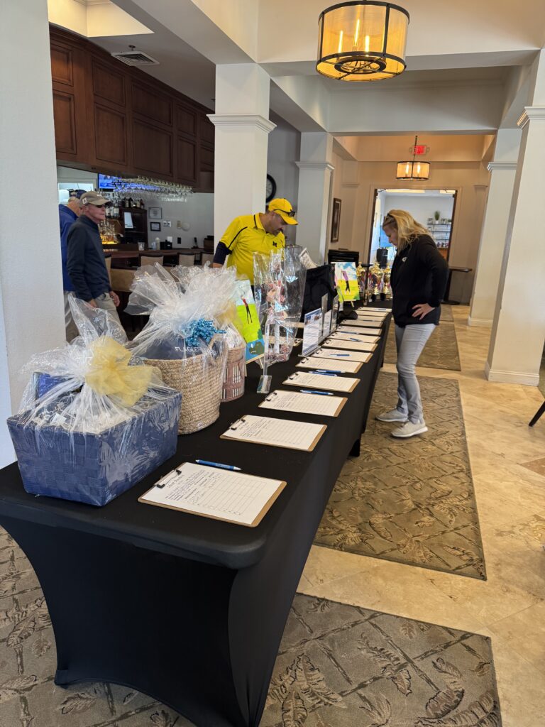 Two people stand at a long table covered with gift baskets and clipboards for a silent auction or raffle in a well lit indoor space with white walls and decorative lighting