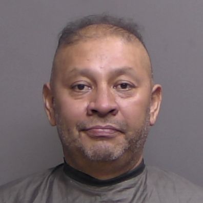 A middle aged man with short, thinning hair and facial stubble is facing the camera against a plain background, wearing a dark, protective garment