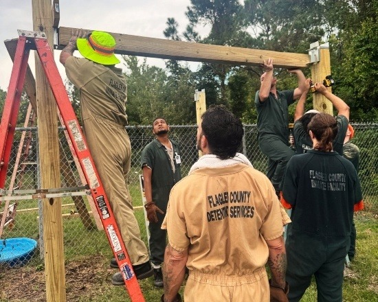 Several people in uniforms labeled "Flagler County Detention Services" work together outdoors to install a wooden beam on a structure near a chain link fence One person is on a ladder, while others support the beam