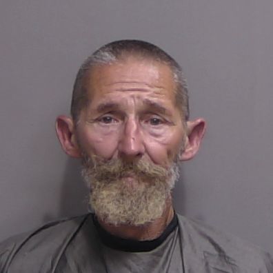 A middle aged man with short hair and a full, bushy beard and mustache, wearing a gray drape, poses for a mugshot against a plain, gray background