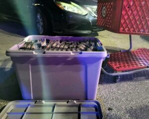 A large plastic bin filled with what appears to be several pairs of sunglasses sits on the ground near a red shopping cart and a parked car at night