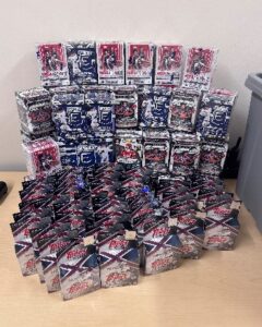 Multiple stacks of boxed trading card packs, including boxes labeled NFL and NBA, are arranged neatly on a table against a plain wall A gray bin sits to the right