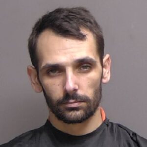 A man with short dark hair, a trimmed beard, and a serious expression wears a dark collared garment in front of a plain gray background