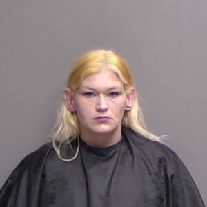 A person with long, straight blond hair wearing a black cape poses against a plain gray background for a mugshot photo